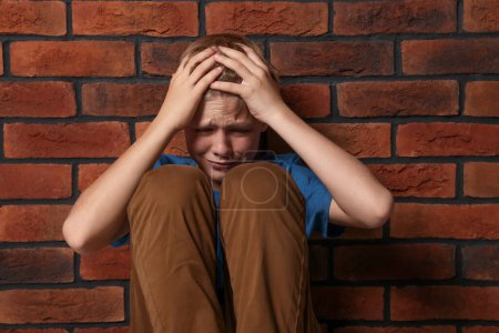 Téléchargez les photos : Un garçon énervé assis près d'un mur de briques. L'intimidation des enfants - en image libre de droit