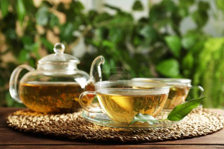 Frischer grüner Tee in Glasbechern mit Untertassen, Blättern und Teekanne auf Holztisch