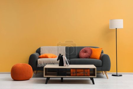 Photo for Stylish grey sofa with colorful pillows, wooden table and lamp near pale orange wall indoors. Interior design - Royalty Free Image