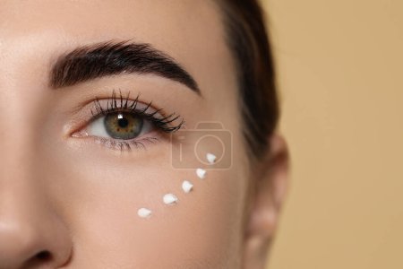 Téléchargez les photos : Gros plan de femme avec produit cosmétique autour des yeux sur fond beige, espace pour le texte. Soins de la peau - en image libre de droit