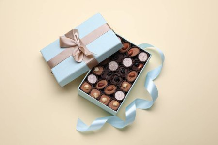 Photo for Open box of delicious chocolate candies and light blue ribbon on beige background, flat lay - Royalty Free Image