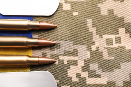 Military ID tags, bullets and Ukrainian flag on pixel camouflage, flat lay. Space for text