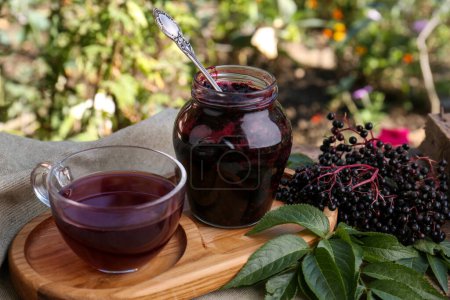 Photo for Elderberry jam, glass cup of tea and Sambucus berries on table outdoors - Royalty Free Image