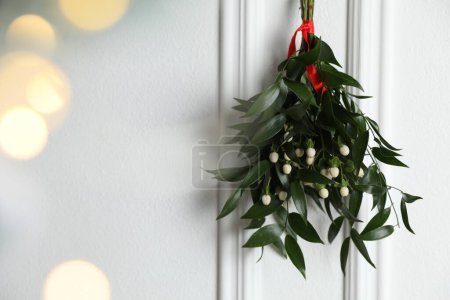 Mistletoe bunch with red ribbon hanging on light background, space for text. Bokeh effect