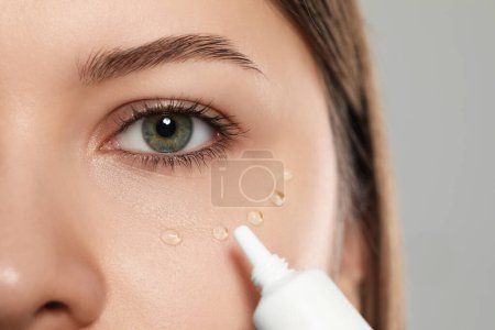 Téléchargez les photos : Jeune femme appliquant de la crème sous les yeux sur fond gris clair, gros plan - en image libre de droit