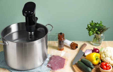 Meat in vacuum packing and other ingredients near pot with sous vide cooker on wooden table. Thermal immersion circulator