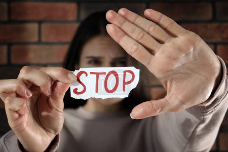 Téléchargez les photos : Concept de violence domestique. Femme tenant du papier avec mot écrit Stop près du mur de briques, se concentrer sur les mains - en image libre de droit