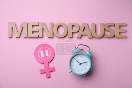 Mot Ménopause en lettres de bois, signe de genre féminin et réveil sur fond rose, pose plate