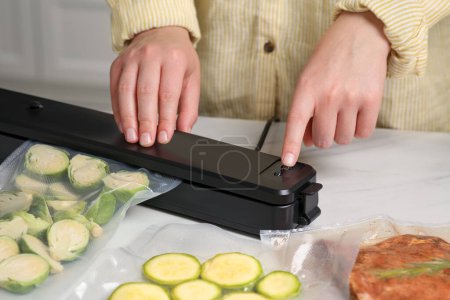 Photo for Woman using sealer for vacuum packing with plastic bag of zucchini at white table, closeup - Royalty Free Image