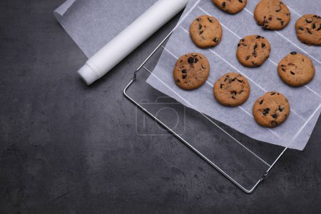 Téléchargez les photos : Rouleau de papier sulfurisé et support de refroidissement avec de savoureux biscuits sur table gris foncé, plat. Espace pour le texte - en image libre de droit
