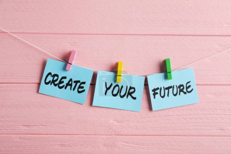 Paper notes with motivational phrase Create Your Future hanging on pink wooden wall