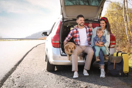 Foto de Los padres, su hija y su perro sentados en el maletero del coche cerca de la carretera, espacio para el texto. Familia viajando con mascota - Imagen libre de derechos
