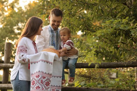 Foto de Feliz linda familia en camisas ucranianas bordadas con pan korovai cerca de la cerca rústica al aire libre. Espacio para texto - Imagen libre de derechos