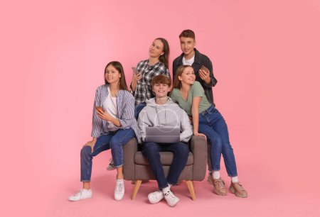 Téléchargez les photos : Groupe d'adolescents heureux utilisant un ordinateur portable et smartphones sur fond rose clair - en image libre de droit