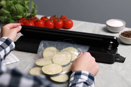 Photo for Woman packing cut eggplant into plastic bag using vacuum sealer on light grey marble table, closeup - Royalty Free Image