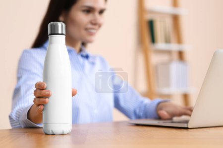 Young woman taking thermo bottle at workplace indoors, focus on hand