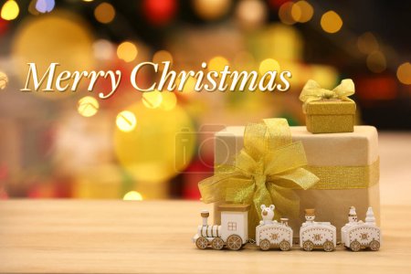 Téléchargez les photos : Carte de voeux avec phrase Joyeux Noël. Belles boîtes-cadeaux avec train de jouets sur table en bois contre les lumières floues festives - en image libre de droit