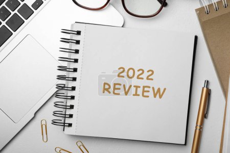 Texto 2022 Revisión escrita en cuaderno, portátil, pluma y vasos en la mesa blanca, la puesta plana
