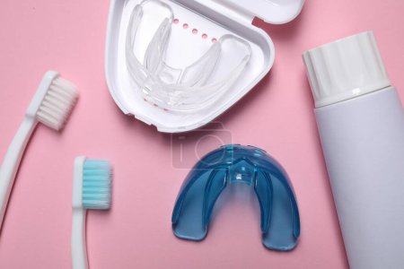 Bite correction. Toothpaste, brushes and dental mouth guards on pink background, flat lay