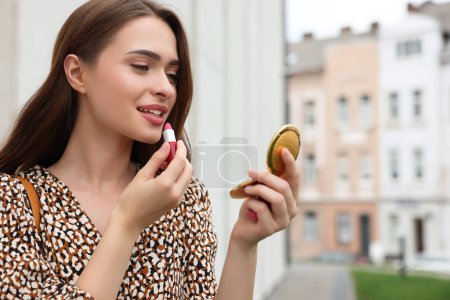 Téléchargez les photos : Belle jeune femme avec miroir de poche cosmétique appliquant du rouge à lèvres à l'extérieur, espace pour le texte - en image libre de droit