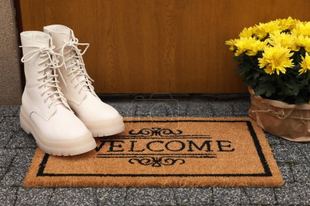 Foto de Doormat with word Welcome, stylish boots and beautiful flowers on floor near entrance - Imagen libre de derechos