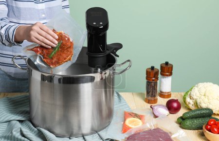 Foto de Woman putting vacuum packed meat into pot with sous vide cooker at wooden table, closeup. Thermal immersion circulator - Imagen libre de derechos
