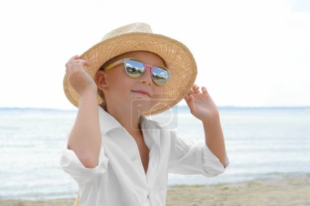 Téléchargez les photos : Petite fille portant des lunettes de soleil et chapeau à la plage par une journée ensoleillée - en image libre de droit