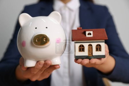 Woman holding piggy bank and little house model on light grey background, closeup
