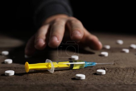 Addicted man reaching to drugs at wooden table, focus on syringe