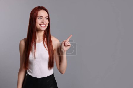 Happy woman with red dyed hair pointing somewhere on light gray background, space for text