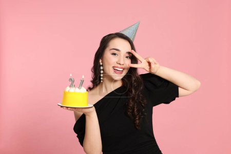 Téléchargez les photos : Coming of age party - 21st birthday. Smiling woman showing peace sign and holding delicious cake with number shaped candles on pink background - en image libre de droit