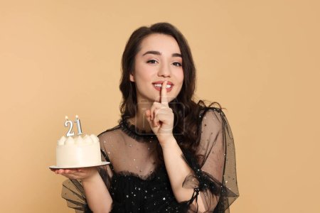 Foto de Coming of age party - 21st birthday. Smiling woman showing silence gesture and holding delicious cake with number shaped candles on beige background - Imagen libre de derechos