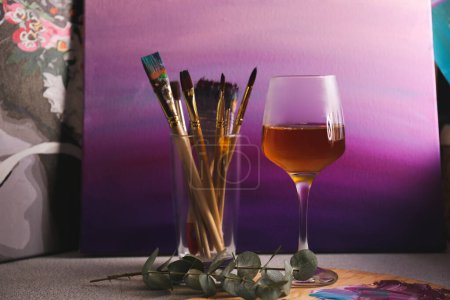 Photo for Glass of tasty wine, brushes with colorful paints and gradient canvas on light gray table - Royalty Free Image