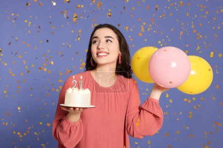 Coming of age party - 21st birthday. Smiling woman holding delicious cake with number shaped candles and balloons and looking at falling confetti on violet background