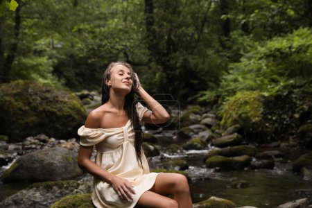Beautiful young woman sitting on rock near mountain river in forest. Space for text