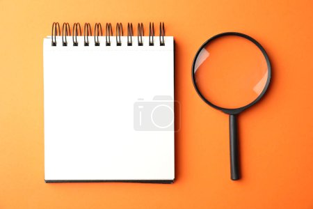 Lupe und leeres Notizbuch auf orangefarbenem Hintergrund, flache Lage mit Platz für Text. Suchbegriff Konzept