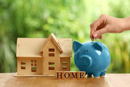 Woman putting money into piggy bank, house model and word Home made of cubes on wooden table outdoors, closeup