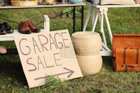 Sign Garage sale written on cardboard near tables with different stuff outdoors