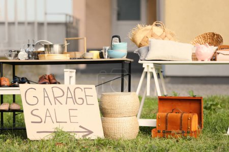 Sign Garage sale written on cardboard near tables with different stuff in yard