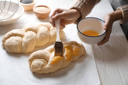 Woman spreading egg yolk over braided dough at white wooden table in kitchen, closeup. Cooking traditional Shabbat challah