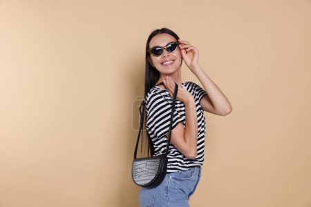 Foto de Beautiful young woman with stylish bag in sunglasses on beige background - Imagen libre de derechos