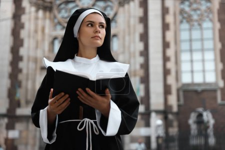 Téléchargez les photos : Young nun reading Bible near cathedral outdoors. Space for text - en image libre de droit