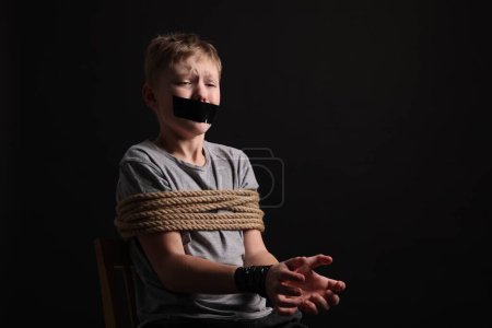 Photo for Little boy with taped mouth tied up and taken hostage against dark background. Space for text - Royalty Free Image