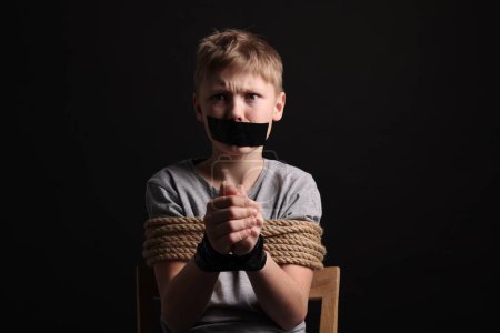 Foto de Little boy with taped mouth tied up and taken hostage against dark background - Imagen libre de derechos
