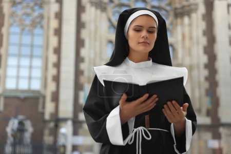 Téléchargez les photos : Young nun reading Bible near cathedral outdoors. Space for text - en image libre de droit