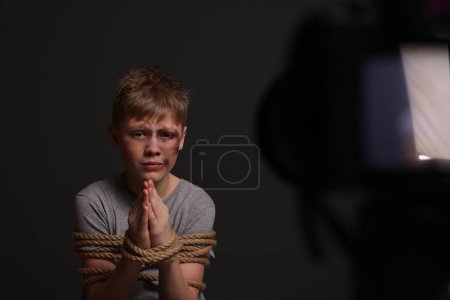 Foto de Little boy with bruises tied up and taken hostage near camera on dark background, selective focus - Imagen libre de derechos
