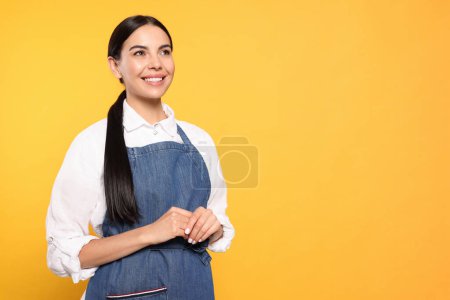 Foto de Young woman in blue jeans apron on yellow background, space for text - Imagen libre de derechos