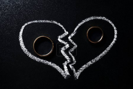Divorce concept. Wedding rings and broken heart drawn on blackboard, flat lay