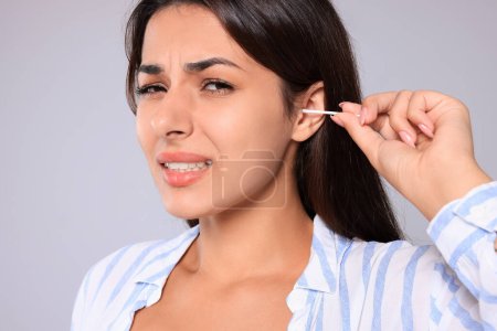 Téléchargez les photos : Young woman cleaning ear with cotton swab on light grey background, closeup - en image libre de droit