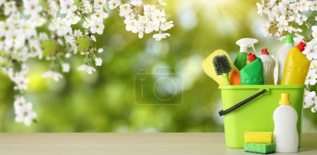 Spring cleaning. Bucket with detergents and tools on wooden surface under blossoming tree against blurred green background, space for text. Banner design